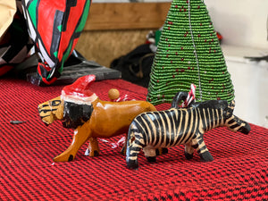 Her Best Foot Forward’s Wooden Holiday Friends—handcrafted Christmas ornaments from Tanzania—feature a lion in a Santa hat and a zebra, displayed on red and black cloth beside a small green Christmas tree decoration.