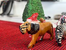 Load image into Gallery viewer, Her Best Foot Forward’s Wooden Holiday Friends—handcrafted Christmas ornaments from Tanzania—include a wooden lion in African style with a red Santa hat on a red checkered surface, plus a sequined green tree and zebra figurine in the background.