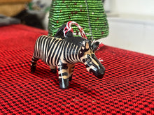 Load image into Gallery viewer, A Wooden Holiday Friends ornament by Her Best Foot Forward, handcrafted in Tanzania, hangs on a red and black patterned surface as a green beaded object appears blurred in the background.