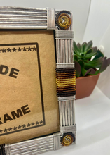 Load image into Gallery viewer, A close-up of the Picha Handmade Beaded Photo Frame by Her Best Foot Forward, crafted with wire, glass, and beads by East African women artisans, in gold, black, and brown hues beside a small potted succulent on a white surface.