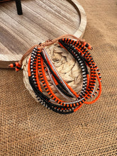 Load image into Gallery viewer, The Shanga Slide Beaded Bracelet by Her Best Foot Forward features black, orange, and silver beads inspired by Maasai jewelry. It rests on a woven coaster with a wooden tray nearby, capturing East African artistry.
