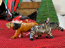 Load image into Gallery viewer, Her Best Foot Forward’s Wooden Holiday Friends—handcrafted lion and zebra ornaments from Tanzania—stand on a red patterned cloth by a green, tree-shaped decoration, highlighting unique African artistry.