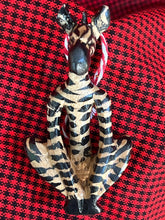 Load image into Gallery viewer, The Wooden Holiday Friends ornament by Her Best Foot Forward features a small, hand-painted figurine with black and tan zebra stripes, sitting cross-legged on a red and black checkered background—a showcase of Tanzanian craftsmanship.
