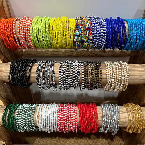 The SIXstax set from Her Best Foot Forward features six colorful, hand-beaded stretch bracelets, neatly arranged by hue—orange, green, yellow, blue, and more—on a wooden rack. Ideal for mixing and stacking your own unique bracelet style.