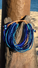 Load image into Gallery viewer, The Shanga Slide Beaded Bracelet by Her Best Foot Forward, inspired by Maasai jewelry from East Africa, features blue and turquoise beads and is shown draped on a weathered tree trunk with sunlit sand and water in the blurred background.