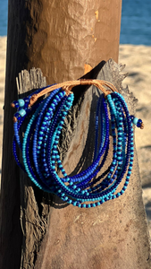 The Shanga Slide Beaded Bracelet by Her Best Foot Forward, inspired by Maasai jewelry from East Africa, features blue and turquoise beads and is shown draped on a weathered tree trunk with sunlit sand and water in the blurred background.