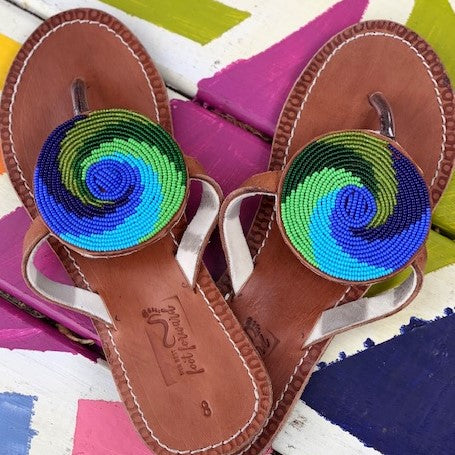 Her Best Foot Forwards Ocean Wave Handmade Beaded Sandals feature brown leather and Maasai beadwork in blue, green, black, and turquoise swirls. This ethical footwear is displayed on a vibrant geometric-patterned surface.
