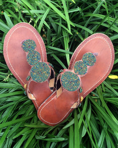 Pools of Green Handmade Beaded Sandals by Her Best Foot Forward feature green and blue Maasai beadwork on brown leather straps, offering nature-inspired, ethical footwear that stands out on lush grass.