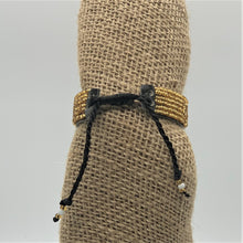 Load image into Gallery viewer, The Her Best Foot Forward Share the LOVE Beaded Bracelet—handmade Maasai jewelry from East Africa—features gold and black beads, an adjustable black cord, and decorative ends, making it a meaningful gift displayed on burlap fabric.