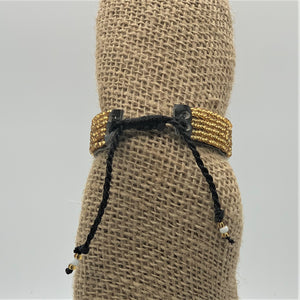 The Her Best Foot Forward Share the LOVE Beaded Bracelet—handmade Maasai jewelry from East Africa—features gold and black beads, an adjustable black cord, and decorative ends, making it a meaningful gift displayed on burlap fabric.