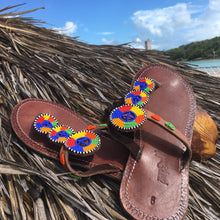 Load image into Gallery viewer, Her Best Foot Forward’s Kelly Handmade Beaded Sandals—sophisticated, ethical footwear adorned with vibrant Maasai beadwork—rest on a thatched surface under blue sky and clouds by the ocean, perfect for summer style.