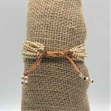 Load image into Gallery viewer, The Shanga Slide Beaded Bracelet by Her Best Foot Forward is an easy slip-on Maasai-inspired piece, with white and gold beads and adjustable brown cords, shown tied around burlap against a plain background.