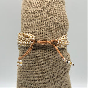 The Shanga Slide Beaded Bracelet by Her Best Foot Forward is an easy slip-on Maasai-inspired piece, with white and gold beads and adjustable brown cords, shown tied around burlap against a plain background.