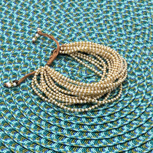 Load image into Gallery viewer, The Shanga Slide Beaded Bracelet by Her Best Foot Forward, inspired by Maasai jewelry, features thin cream and white beads with a brown string clasp, arranged on a blue and green woven mat.