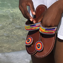 Load image into Gallery viewer, A person holds Her Best Foot Forward’s Im With the Band Handmade Beaded Sandals—vibrant rainbow ethical footwear with Maasai beadwork—while standing by clear, shallow water and wearing white shorts.