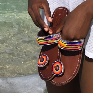 A person holds Her Best Foot Forward’s Im With the Band Handmade Beaded Sandals—vibrant rainbow ethical footwear with Maasai beadwork—while standing by clear, shallow water and wearing white shorts.
