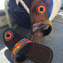 Load image into Gallery viewer, Her Best Foot Forward’s Im With the Band Handmade Beaded Sandals—featuring vibrant rainbow beadwork—rest on a gray surface near a large blue-and-white buoy with rope attachment.