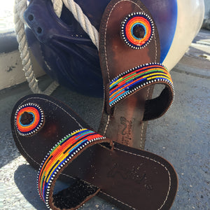 Her Best Foot Forward’s Im With the Band Handmade Beaded Sandals—featuring vibrant rainbow beadwork—rest on a gray surface near a large blue-and-white buoy with rope attachment.