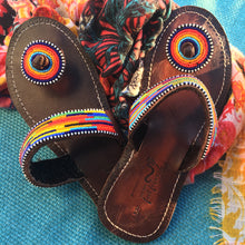 Load image into Gallery viewer, Her Best Foot Forward’s “I’m With the Band” Handmade Beaded Sandals—vibrant rainbow ethical footwear—rest on a bright blue woven surface, with a hint of floral fabric nearby.