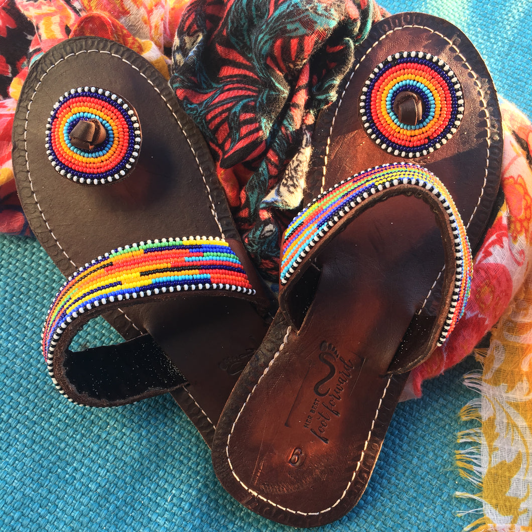 Her Best Foot Forward’s “I’m With the Band” Handmade Beaded Sandals—vibrant rainbow ethical footwear—rest on a bright blue woven surface, with a hint of floral fabric nearby.