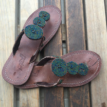 Load image into Gallery viewer, Her Best Foot Forwards Pools of Green Handmade Beaded Sandals feature intricate Maasai beadwork in blue, green, yellow, and black on brown leather, creating unique nature-inspired ethical footwear displayed on a wooden surface.