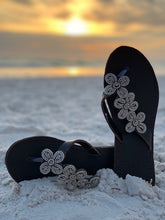 Load image into Gallery viewer, Her Best Foot Forwards Addison Handmade Beaded Sandals, featuring Maasai beadwork, sit on white sand as a sunset blurs over the ocean in the background—ethical summer footwear with floral accents.