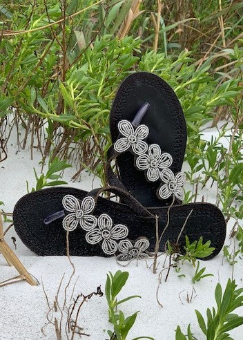 The Addison Handmade Beaded Sandals by Her Best Foot Forward feature Maasai beadwork, offering ethical summer footwear that stands out in black with white floral patterns on white sand and green grass.