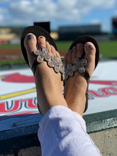 Load image into Gallery viewer, Relaxing with feet up in white pants, she wears Her Best Foot Forwards Addison Handmade Beaded Sandals—ethical summer footwear featuring Maasai beadwork—as a blurred outdoor stadium and colorful logo appear in the background.
