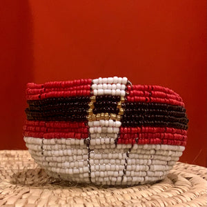 A beaded bracelet with a red, white, black, and gold Poké Ball pattern sits on a woven surface paired with Her Best Foot Forward Santa Baskets—handcrafted Christmas baskets from Tanzania—to create the perfect holiday gift set.