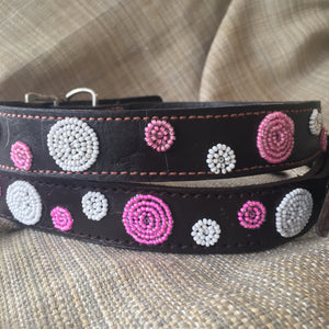A close-up of two Petunia Pink Beaded Dog Collars by Her Best Foot Forward, handmade in East Africa, showcases pink and white bead patterns on dark leather, resting on textured beige fabric.