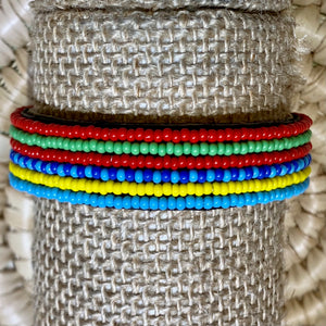 The Safari Stripes Beaded Bracelet by Her Best Foot Forward features bold, alternating stripes inspired by Maasai colors. Handmade in East Africa, each bracelet showcases uniform beads and is displayed on a textured beige stand.