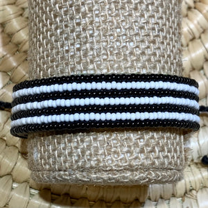 The Safari Stripes Beaded Bracelet by Her Best Foot Forward is a handmade Maasai-inspired accessory from East Africa, featuring alternating black and white beaded stripes, beautifully presented on a woven beige surface.