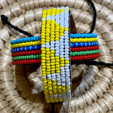 Load image into Gallery viewer, The Safari Stripes Beaded Bracelet by Her Best Foot Forward is a handmade Maasai-inspired accessory featuring alternating colorful stripes, displayed on a woven straw mat.