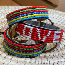 Load image into Gallery viewer, Three beaded bracelets are shown: two with rainbow stripes and one Her Best Foot Forward “Share the LOVE Beaded Bracelet” from East Africa, handmade Maasai jewelry with LOVE in white beads, all displayed on a woven surface.
