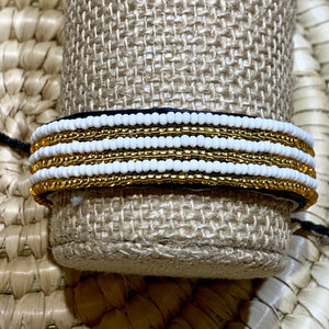 The Safari Stripes Beaded Bracelet by Her Best Foot Forward features handmade Maasai jewelry with alternating white and gold stripes, beautifully displayed on a burlap roll atop a woven mat.