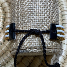 Load image into Gallery viewer, A close-up of Her Best Foot Forwards Safari Stripes Beaded Bracelet, a handmade Maasai jewelry piece featuring alternating white and gold beads on black tabs with a black cord, set against a woven straw background.