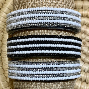 The Safari Stripes Beaded Bracelet by Her Best Foot Forward is handmade Maasai jewelry from East Africa featuring alternating stripes of white, black, and silver beads, beautifully displayed on a woven surface.