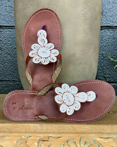 The Mary Handmade Beaded Sandals by Her Best Foot Forward feature brown leather, white floral Maasai beadwork on the straps, and are displayed on wood with a black and beige background. Timeless, ethical footwear.