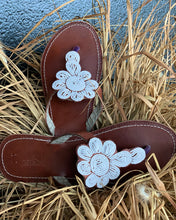 Load image into Gallery viewer, Her Best Foot Forwards Mary Handmade Beaded Sandals feature brown leather and Maasai beadwork with white floral designs on the straps, blending timeless ethics and style. Displayed on dry grass.