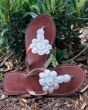Load image into Gallery viewer, The Mary Handmade Beaded Sandals by Her Best Foot Forward feature brown straps adorned with white floral Maasai beadwork, shown outdoors on mulch with green leafy plants in the background. Timeless, ethical footwear.