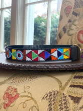 Load image into Gallery viewer, The Tapestry Beaded Dog Collar by Her Best Foot Forward displays an intricate handmade Maasai pattern, resting on leather with patterned upholstery and greenery outside, showcasing ethical East African craftsmanship.