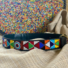 Load image into Gallery viewer, A black leather Tapestry Beaded Dog Collar by Her Best Foot Forward, featuring Maasai beadwork, rests on beige fabric. In the background, a vibrant multicolored collar showcases its intricate handmade ethical design from East Africa.