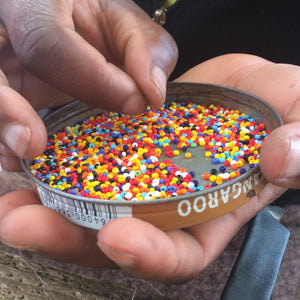 A person sorts colorful beads like those on Her Best Foot Forwards Kelly Handmade Beaded Sandals, featuring Maasai beadwork; the metal lid they use has a barcode and text around its edge.