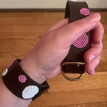 Load image into Gallery viewer, A hand holds the Petunia Pink Beaded Dog Collar by Her Best Foot Forward—an ethical, handmade Maasai accessory with spiral beadwork, wide dark leather, brass D-ring and buckle—displayed on a wooden floor.