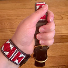 Load image into Gallery viewer, A hand displays Her Best Foot Forwards Red Hot Dog Beaded Dog Collar—an ethical, handmade Maasai pet accessory from East Africa—with a matching beaded bracelet, set against a wooden floor.