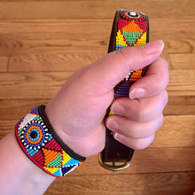Load image into Gallery viewer, A hand wears a colorful beaded bracelet and holds the Tapestry Beaded Dog Collar by Her Best Foot Forward, showcasing its intricate handmade Maasai ethical design from East Africa and bright geometric patterns against wooden flooring.