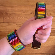 Load image into Gallery viewer, A hand holds the Ethan Almighty Beaded Dog Collar by Her Best Foot Forward, a handmade Maasai ethical pet accessory from East Africa with colorful beadwork and a matching bracelet, displayed above a wooden floor.