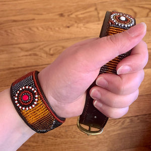 A hand holds the Savanna Beaded Dog Collar by Her Best Foot Forward—an ethical, handmade Maasai pet accessory with vibrant red, white, and orange beadwork—against a wooden floor. Another matching beaded piece wraps the wrist.
