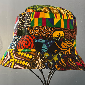 The Ndoo Reversible Bucket Hat by Her Best Foot Forward features vibrant geometric and abstract patterns in red, yellow, green, blue, and black Kitenge fabric from East Africa, displayed on a metal stand against a gray background.