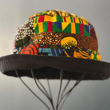 Load image into Gallery viewer, The Ndoo Reversible Bucket Hat by Her Best Foot Forward is handmade from vibrant, geometric-patterned Kitenge fabric from East Africa, shown resting on a wire stand against a plain gray background.
